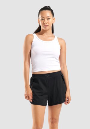 Ung kvinde med hestehale iført en hvid ærmeløs crop top og sorte atletikshorts mod en ensfarvet lys baggrund.