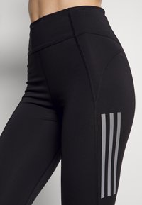 Schwarze Sportleggings mit einem hohen Bund, aus dehnbarem Stoff gefertigt. Mit drei vertikalen grauen Streifen auf dem äußeren Oberschenkel.