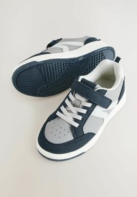 Baskets avec une sangle velcro, présentant un mélange de matériaux bleu marine et gris, tige en textile, semelle en caoutchouc et design de bout perforé.
