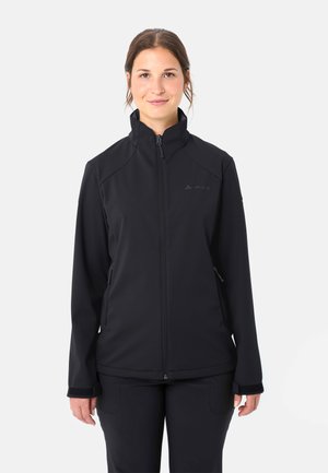 Femme debout, portant une veste noire zippée avec un col montant et des poches latérales zippées, face à l'avant sur un fond uni.