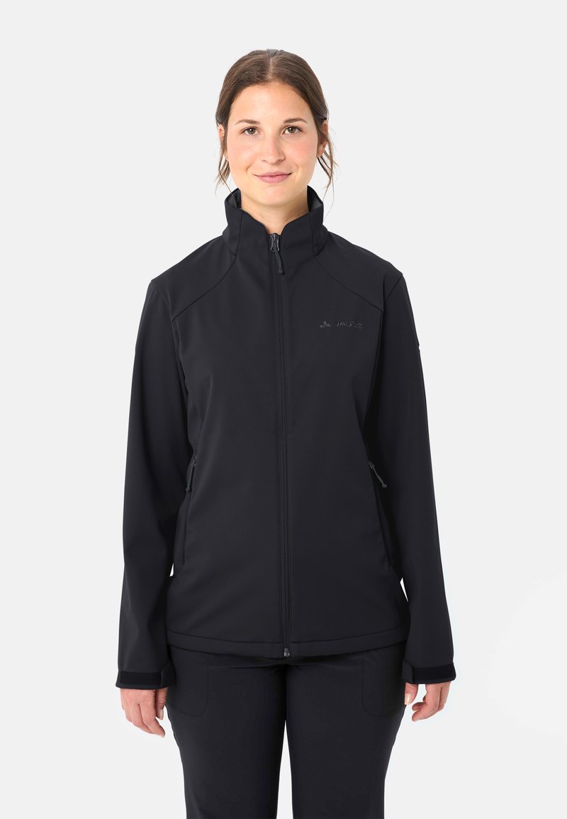 Donna in piedi, indossa una giacca nera con zip, colletto alto e tasche laterali con cerniera, rivolta verso l'obiettivo su uno sfondo semplice.