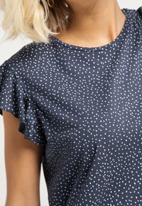 Mörkblå topp med vitt prickmönster, korta volangärmar, rund halsringning och slät textur som är lämplig för avslappnad klädsel.