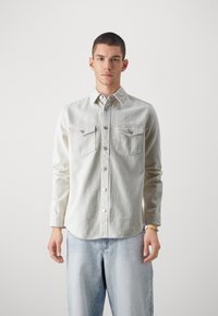 Cămașă din denim bej deschis, cu mâneci lungi, închidere cu nasturi în față, două buzunare pe piept și un tiv ușor curbat. Pur și simplu cu blugi albastru deschis.