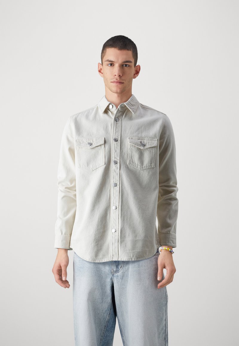 Cămașă din denim bej deschis, cu mâneci lungi, închidere cu nasturi în față, două buzunare pe piept și un tiv ușor curbat. Pur și simplu cu blugi albastru deschis.