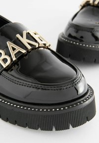 Mocassini in pelle verniciata nera con suola platform chunky, hardware dorato "BAKER" e dettagli laterali testurizzati con borchie.