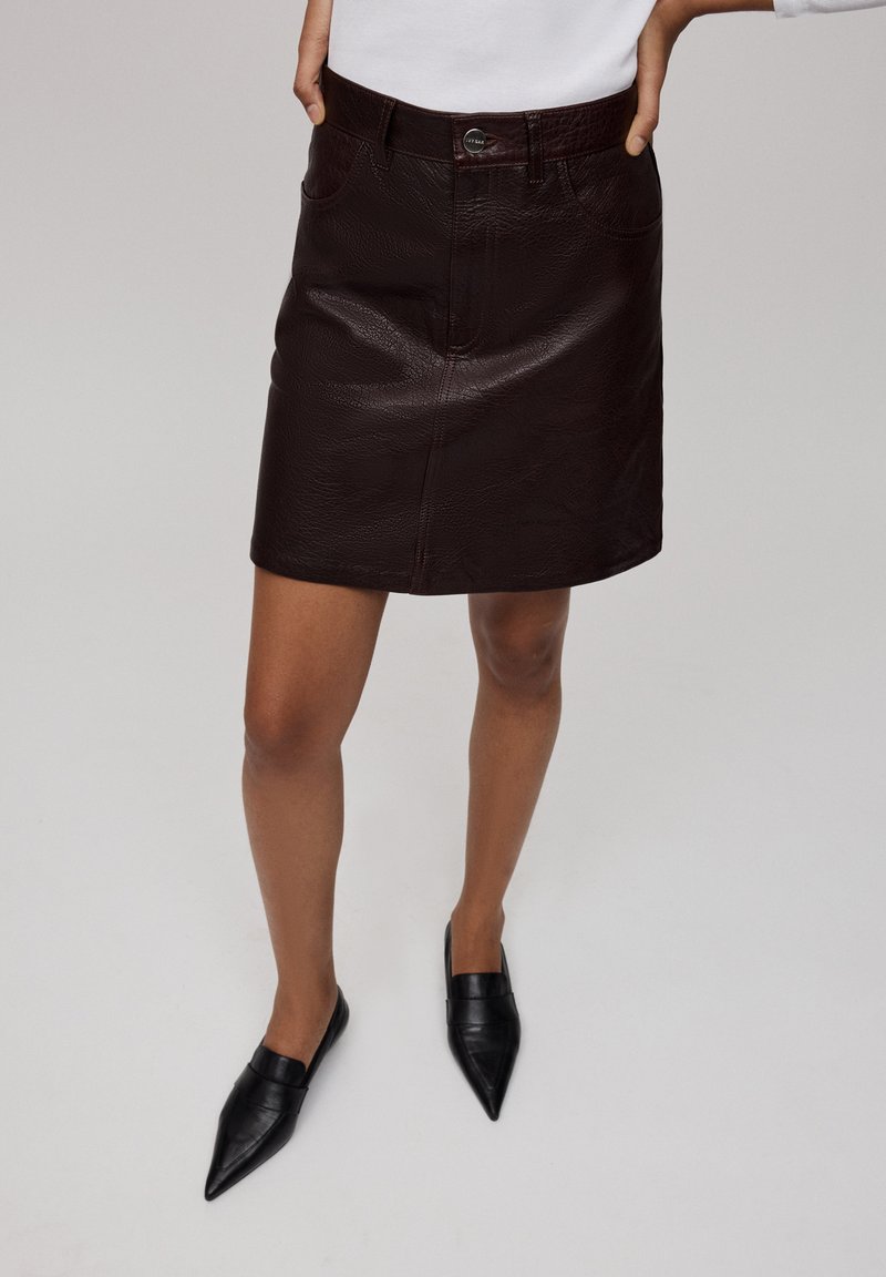 Gonna mini in pelle borgogna con una finitura testurizzata, tasche frontali e chiusura con bottone, abbinata a ballerine nere con punta appuntita.