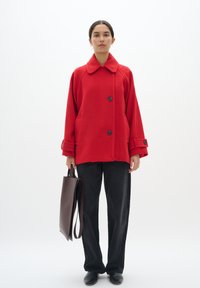 Rode oversized wollen jas met een ronde kraag, voorknopen en twee zakken, gecombineerd met zwarte broeken en een bruine totebag.