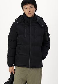 Schwarze Daunenjacke mit hohem Kragen, Reißverschluss vorne, zwei Brusttaschen und abnehmbarer Kapuze. Das Material erscheint glatt mit einem gesteppten Muster.