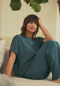 Teal loungewear-setti, joka sisältää väljän lyhythihaisen paidan ja yhteensopivat kapenevat housut, valmistettu teksturoidusta, pehmeästä materiaalista.