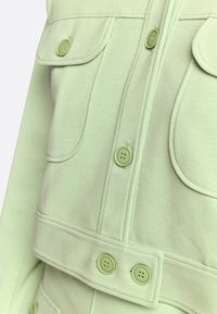 Light green jacket with a soft texture, featuring large flap pockets and matching buttons. The design includes visible stitching details.