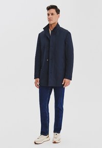 Granatowa, lekka kurtka z wysokim kołnierzem i zapięciem na guziki; zestawiona z ciemnymi dżinsami i kremowymi trampkami. Gładka faktura materiału.