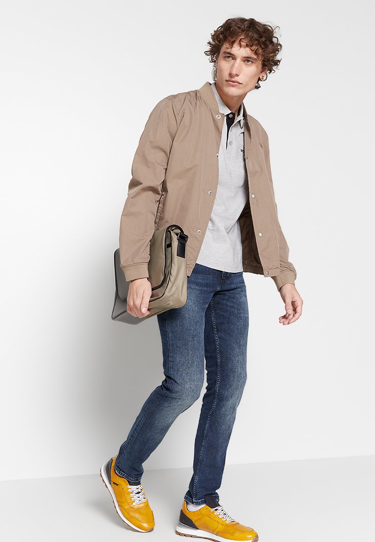 Tan bomberjack over een grijze polo, blauwe jeans en gele sneakers. Houdt een grijze tas vast, wat een casual, gelaagde look laat zien.