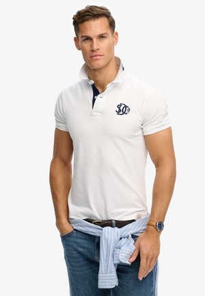 Jeune homme portant un polo blanc avec un logo, un jean bleu, une chemise bleu rayée nouée autour de la taille, et une montre-bracelet au bras gauche.