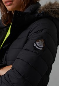 Svart puffjacka med quiltad yta, utrustad med en pälsfodrad huva och en rund, flerfärgad patch med texten "Superdry" på ärmen.
