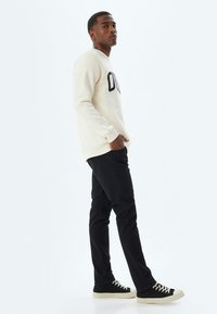 Sudadera de color crema claro con texto negro, combinada con pantalones ajustados negros y zapatillas negras, de pie frente a un fondo blanco liso.