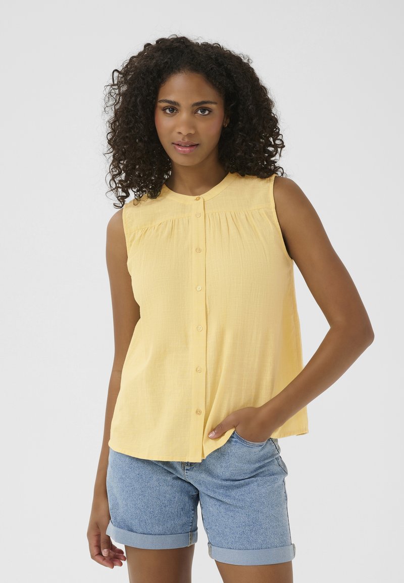 Chemise sans manches jaune avec une finition texturée, fermeture par boutons et détails froncés aux épaules, associée à un short en denim.