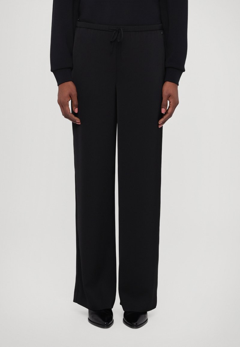 Personne portant un pantalon noir à jambes larges avec une taille à cordon et des chaussures noires à bout pointu, debout devant un fond clair uni.