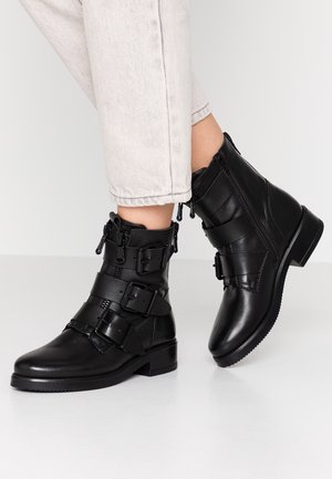 Bottes à chevilles en cuir noir avec trois larges sangles et boucles, fermetures éclair latérales, portées avec un pantalon court de couleur claire sur un fond uni.