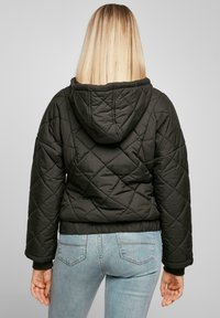 Svart quiltet jakke med hette, med diamantmønster og ribbestrikkede mansjetter, sammen med lyseblå jeans. Utsikt fra baksiden.