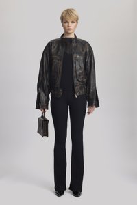 Veste en cuir marron avec poches zippées, pull noir à côtes, et pantalon noir évasé, tenant un petit sac à main marron. Texture lisse.