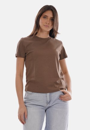 Femme aux cheveux bruns lisses portant un t-shirt marron uni à manches courtes et un jean bleu clair, debout devant un fond blanc.