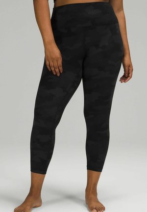 Rond persoon die zwarte camouflage cropped leggings met hoge taille draagt en blootsvoets staat tegen een grijze achtergrond.