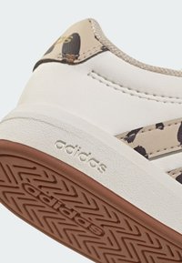 Scarpa da ginnastica in pelle bianca con accenti beige e nero a stampa leopardata. Presenta suola testurizzata con motivo a spina di pesce e logo "adidas" in rilievo.