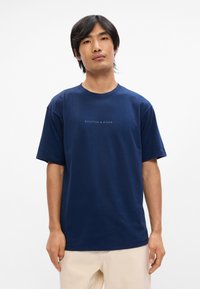 Μπλέ βαμβακερό μπλουζάκι με κοντό μανίκι και στρογγυλή λαιμόκοψη, με το λογότυπο "SCOTCH & SODA" τυπωμένο σε πιο ανοιχτή απόχρωση στο μπροστινό μέρος.