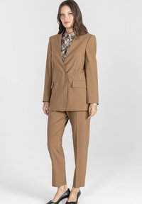Brązowy, dopasowany garnitur z wyraźnie zarysowanym żakietem z kieszeniami, zestawiony z wąskimi spodniami. Nosi się go na wzorzystą koszulę, dopełniony czarnymi butami.