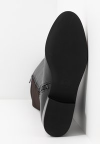 Suela de goma negra con agarre texturizado, parte superior de cuero gris con detalle de cremallera diagonal y acento de tela marrón en el lateral.