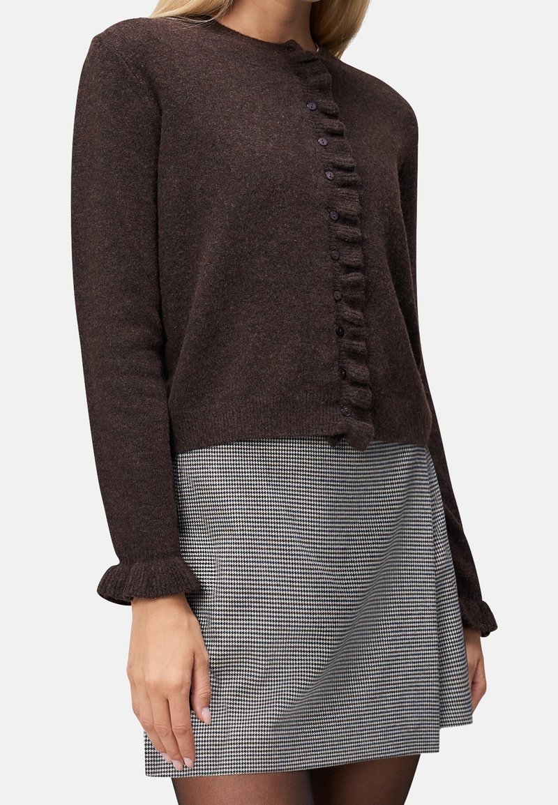 Femme portant un cardigan boutonné à manches longues marron foncé avec des bords volantés et une mini-jupe à motif pied-de-poule noir et blanc avec des collants.