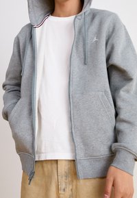 Felpa grigia con zip, dotata di tasca frontale, coulisse regolabili e un piccolo logo sul petto. Indossata sopra una maglietta bianca.