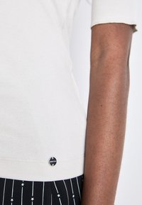 Crème ribbed t-shirt met korte mouwen, voorzien van een ronde zwarte logo-accent nabij de zoom. Draagt een zwarte gestreepte stoffen broek.