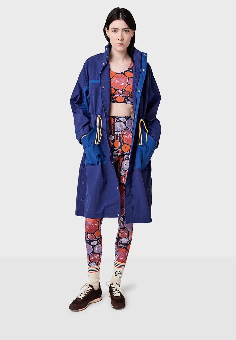 Woman wearing a navy blue long coat over floral crop top and leggings with orange, purple flowers, striped socks, and dark brown sneakers, standing.