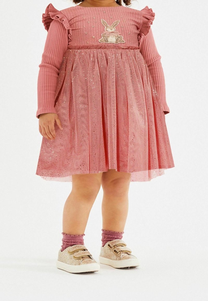 Enfant portant une robe rose à manches longues avec une application de lapin, jupe en tulle pailleté, chaussettes roses et baskets à scratch pailletées, debout sur un sol blanc.