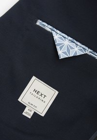 Tissu bleu marine foncé avec une pochette à motifs géométriques blancs et une étiquette blanche Next Tailoring indiquant "Slim Fit 40R Made in Bangladesh."