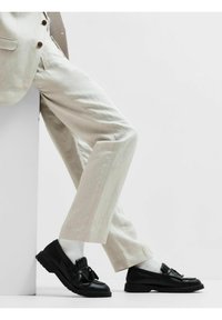 Svarta läderloafers med en rosettdetalj, kombinerat med ljusbeige linbyxor. Strumporna är vita och något strukturerade, synliga ovanför skorna.
