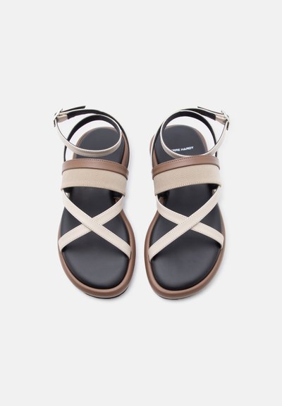 Ζευγάρι επίπεδων σανδαλιών με μαύρες σόλες, καφέ άκρες, μπεζ σταυρωτά και φαρδιά λουριά και κούμπωμα στον αστράγαλο σε λευκό φόντο.