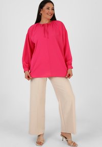 Top lungo a maniche con scollo arricciato e dettaglio con fiocco di un brillante colore rosa, abbinato a pantaloni beige chiaro a gamba larga e tacchi beige con laccetti.