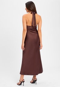 Robe halter en satin marron avec dos nu, comprenant un détail de ruban noué et une silhouette en A longueur sol. Sandales à talons noires visibles.