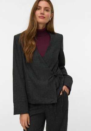 Femme portant un blazer drapé gris foncé et un pantalon assorti sur un col roulé violet, debout devant un fond blanc uni.