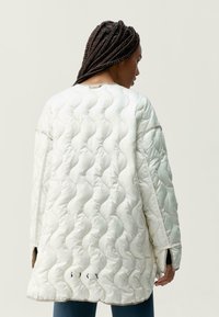 Prošívaná oversized bunda v odstínu off-white s vlnkovaným vzorem, s dlouhými rukávy, bočními rozparky a minimalními hnědými akcenty.