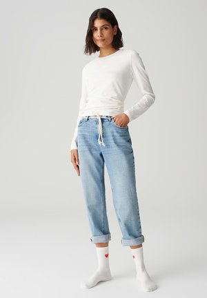 Weißes Langarmoberteil kombiniert mit hellblauen, hoch taillierten Jeans, die hochgekrempelt sind. Das Model trägt weiße Socken mit roten Herzakzenten.