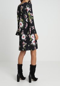 Robe à manches cloche florale avec un fond noir, présentant de grandes fleurs blanches et roses, longueur au-dessus du genou, associée à des bottes à talons hauts noires.