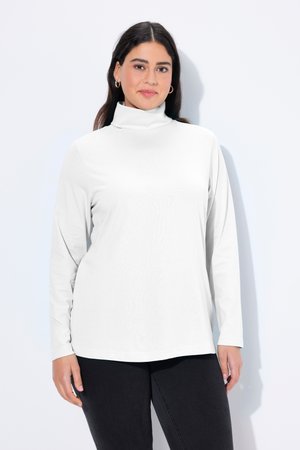Frau mit langen dunklen Haaren, die Creolen trägt, ein weißes Rollkragentop und schwarze Hose, stehend vor einem einfachen hellen Hintergrund.