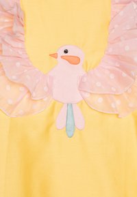 Un tissu jaune présente une application d'oiseau rose avec un bec orange et des pieds bleus. Des volants roses à pois améliorent l'encolure.