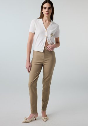 Vrouw staat en draagt een witte korte mouw cardigan met knopen en een gouden embleem, beige aansluitende broek en crème laaggehakte schoenen met strikjes.