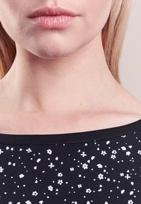 Indumento nero con motivo floreale bianco e macchiato, caratterizzato da una scollatura ampia e un aspetto liscio e testurizzato. Primo piano dell'area del colletto.