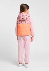 Gilet rembourré en rose clair avec un bas orange ; présente des motifs ludiques. Associé à un jogging rose et des chaussures blanches à enfiler.
