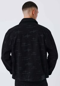 Chaqueta de mezclilla negra con una textura estampada que presenta sutiles motivos de estrellas, detalles cosidos y una etiqueta en el dobladillo inferior.